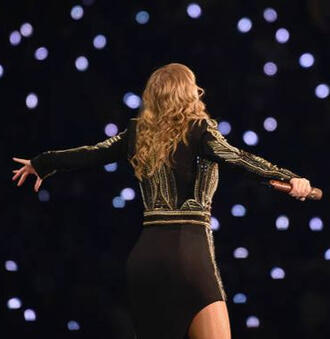 Taylor Swfit de espaldas, en un concierto. El fondo es oscuro con varios puntitos blancos (linternas de los celulares). A Taylor le cae el cabello rubio y ondulado suelto por la espalda, usa un traje negro de manga larga con detalles dorados.